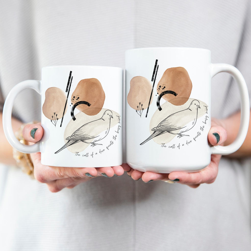  A woman holding two ceramic mugs featuring a sketch of a morning dove against a background of abstract shapes in neutral earth tone colors.